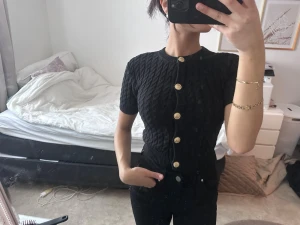 Svart kabelstickad kortärmad kofta - Svart kabelstickad kofta med korta ärmar och snygga guldknappar framtill. Rund halsringning och figurnära passform ger en stilren look. Perfekt att styla med jeans eller kjol för en trendig outfit.