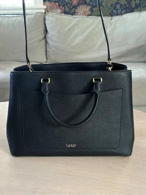 Svart handväska från Lauren Ralph Lauren - Stilren svart handväska i läder från Lauren Ralph Lauren med både handtag och justerbar axelrem. Väskan har guldfärgade metalldetaljer och en klassisk, fyrkantig form. Perfekt för dig som vill ha en snygg och rymlig väska med exklusiv känsla.Väskan är näst intill helt oanvändbar då jag fick den av min moster, men känner att den inte riktigt passar mig
