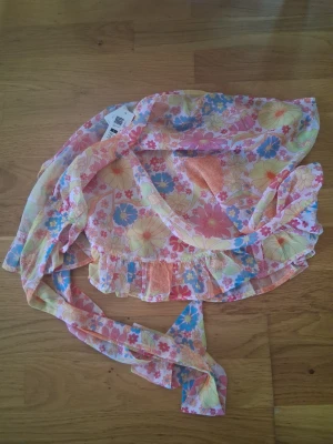 Blommig strandkimono - Färgglad strandkimono från cubus i storlek L. Tunt och luftigt material med blommigt mönster i rosa, orange, blått och gult. Perfekt att slänga över bikinin på stranden. Knytband i midjan och volangkant nedtill för en extra söt look.