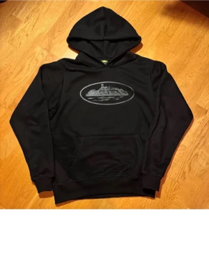 Svart hoodie med tryck av fästning - Svart hoodie med stor huva och magficka. På bröstet finns ett tryck i grått av en fästning eller borg inuti en oval ram. Hoodien har ribbade muddar vid ärmslut och nederkant. Perfekt för en chill och avslappnad stil.