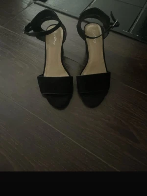 Svarta pumps med öppen tå - Stilrena svarta pumps från Nelly med öppen tå och bred rem över foten. Skorna har en justerbar ankelrem och klack, vilket ger en elegant siluett. Materialet ser ut att vara mockaimitation och innersulan är ljusbeige.
