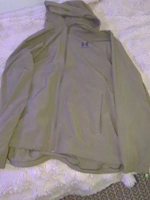 Olivgrön vindjacka från Under Armour - Snygg olivgrön vindjacka från Under Armour med huva och dragkedja framtill. Jackan har två sidofickor och är tillverkad i ett lätt, vattenavvisande material. Perfekt för blåsiga dagar och sportiga outfits.
