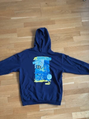 Blå Manchester City hoodie Puma - Snygg blå hoodie från Puma med Manchester City-tryck i blått, gult och vitt på ryggen. Hoodien har huva och ribbade muddar vid ärmslut och nederkant. Perfekt för dig som älskar fotboll och vill ha en unik streetwear-look.