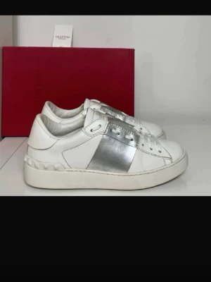 Valentino vita sneakers med silverdetalj - Snygga vita sneakers från Valentino med en bred silverfärgad remsa på sidan. Skorna har klassisk låg modell, vita snören och diskreta nitar på hälen. Tillverkade i läder för en lyxig känsla och stilren look.