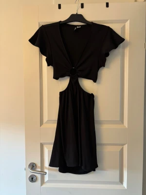Svart cut out klänning från H&M Divided - Svart kortklänning från H&M Divided med cut out-detalj i midjan och ring i mitten. Klänningen har korta ärmar och v-ringad hals. Perfekt för dig som vill ha en trendig och edgy look.