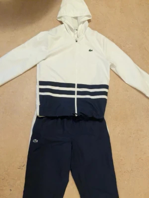 Lacoste Sport vit och marinblå träningsdress - Snygg träningsdress från Lacoste Sport i vitt och marinblått. Jackan har huva, dragkedja och två horisontella ränder över magen. Byxorna är marinblå med vit detalj på sidorna. Båda delarna har Lacostes klassiska krokodillogga. Perfekt för sport eller chill.
