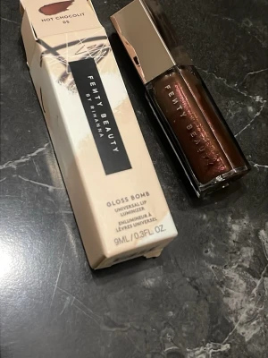 Fenty Beauty Gloss Bomb Hot Chocolit - Fenty Beauty Gloss Bomb i nyansen Hot Chocolit är ett läppglans med brun, skimrande färg. Kommer i en snygg, sexkantig förpackning med guldigt lock. Ger glansiga och fylliga läppar med en hint av chokladton.