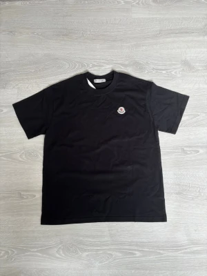 Svart Moncler t-shirt  - Snygg svart t-shirt från Moncler med klassisk passform och rund hals. T-shirten har ett diskret Moncler-logomärke på bröstet och är tillverkad i mjuk bomull. Perfekt för en clean och stilren look.