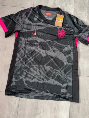 Nike svart fotbollströja med mönster - Snygg svart fotbollströja från Nike med grått grafiskt mönster och rosa detaljer. Tröjan har korta ärmar, rund hals och en rosa Nike-logga samt ett lejonmärke på bröstet. Tillverkad i lätt och ventilerande funktionsmaterial.