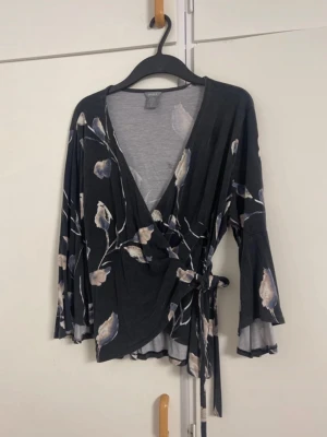 Blommig omlottopp från Vero Moda - Snygg svart omlottopp från Vero Moda med blommigt mönster i beige och blå toner. Toppen har trekvartsärmar med lätt utsvängd form och knyt i midjan för en feminin siluett. Perfekt för dig som gillar en chic och avslappnad stil.