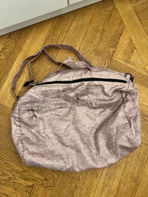 Ljuslila mjuk resväska med axelrem - Säljer en ljuslila resväska i mjukt tyg med subtilt mönster. Väskan har både axelrem och handtag, samt dragkedjefack på framsidan. Perfekt för weekendresor eller gymmet, lätt att bära och packa.
