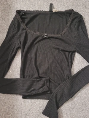 Svart ribbad långärmad topp - Snygg svart ribbad topp med lång ärm och spetsdetalj längs urringningen. Liten rosett framtill ger en söt touch. Perfekt till jeans eller kjol för en trendig look.