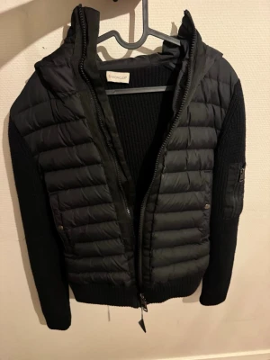 Svart pufferjacka från Moncler - Säljer en svart cardigan från Moncler med stickade ärmar och quiltad front. Jackan har huva, dragkedja framtill och en ficka med dragkedja på ena ärmen. 