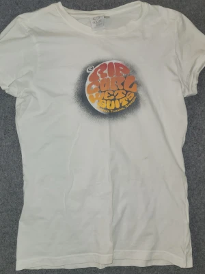 Vit Rip Curl t-shirt med tryck - Säljer en vit t-shirt från Rip Curl med färgglatt retrotryck i orange och gul på bröstet. T-shirten har rund hals och korta ärmar. Perfekt för dig som gillar surfvibbar och streetstyle.