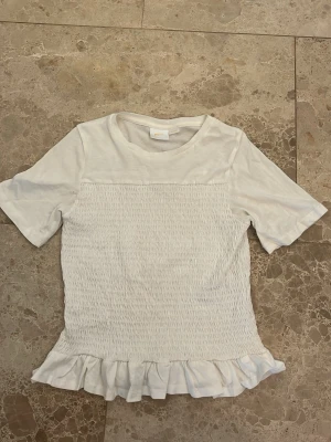 Vit topp med volangkant och rynk - Söt vit topp med korta ärmar och rund hals. Toppen har rynkdetaljer över hela kroppen och en volangkant längst ner som ger en feminin touch. Tillverkad i mjukt bomullsmaterial, perfekt för en avslappnad men ändå stilsäker look.