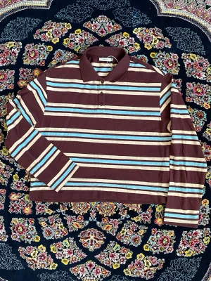 Brun randig långärmad polotopp - Säljer en brun långärmad polotopp med breda horisontella ränder i vitt, blått och beige. Toppen har krage och knäppning framtill. Perfekt för dig som gillar retrovibbar och vill ha en bekväm och stilren look.