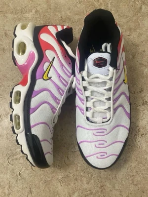 Nike Air Max Plus vita/lila sneakers - Nike Air Max Plus sneakers med vit mesh, lila och rosa vågiga detaljer, gula Swoosh-loggor och svarta samt röda inslag. Synliga Air Max-enheter i sulan och klassisk TN-logga på plösen. Perfekta för dig som gillar färgstarka och unika sneakers.