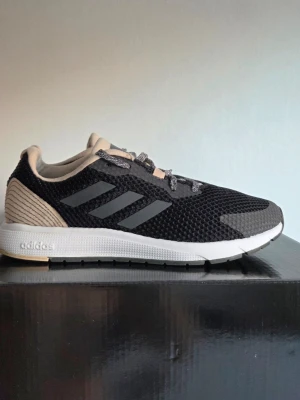 Svarta Adidas sneakers med beige detaljer - Snygga svarta Adidas Sooraj Running sneakers med mesh-överdel, beige häl och plös samt klassiska tre ränder på sidan. Skorna har gråa sulor och grå detaljer vid tån. Perfekta för en sportig och avslappnad stil. 