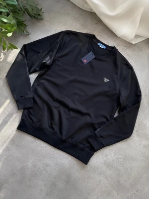 Svart sweatshirt från Prada - Stilren svart sweatshirt från Prada med rund halsringning och diskret trekantslogga på bröstet. Tröjan har långa ärmar, ribbade muddar och är tillverkad i ett mjukt material som ger en clean och modern look.