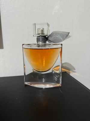 Lancôme La Vie Est Belle parfym 50ml - Säljer en elegant parfym från Lancôme, La Vie Est Belle, i en genomskinlig glasflaska med orangefärgad vätska. Flaskan har en fyrkantig form och ett dekorativt grått band runt halsen. Perfekt för dig som gillar lyxiga dofter.