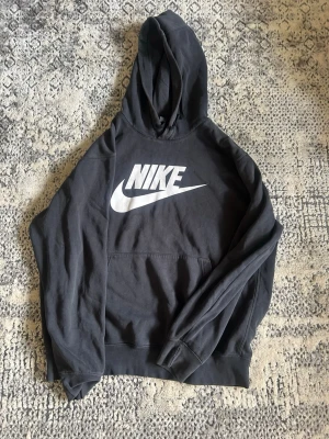 Svart Nike hoodie med stor logga - Snygg svart hoodie från Nike med stor vit logga och swoosh på bröstet. Klassisk känguruficka framtill och mjukt material på insidan. Perfekt för en avslappnad och sportig stil. Huva med dragsko för extra komfort.