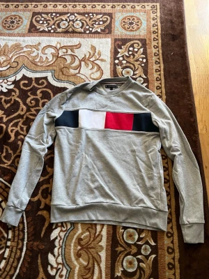 Grå sweatshirt Tommy Hilfiger - Snygg grå sweatshirt från Tommy Hilfiger med klassiskt färgblocksmönster i marinblått, vitt och rött över bröstet. Tröjan har rund halsringning och ribbade muddar vid ärmslut och nederkant. Perfekt för en avslappnad och trendig look.