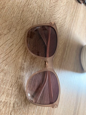 Beige solglasögon från Guess - Snygga solglasögon från Guess med stora, fyrkantiga bågar i beige plast och bruna tonade glas. Sidorna har en metallisk detalj med Guess-loggan. Perfekta för soliga dagar och ger en cool vibe till din outfit.