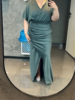 Grön omlott långklänning i satin - Elegant långklänning i glansigt grönt satintyg med omlott överdel och v-ringning. Klänningen har korta, vida ärmar och markerad midja med drapering samt en slits framtill. Perfekt för dig som vill ha en stilren och lyxig look. Pris kan diskuteras!🌸