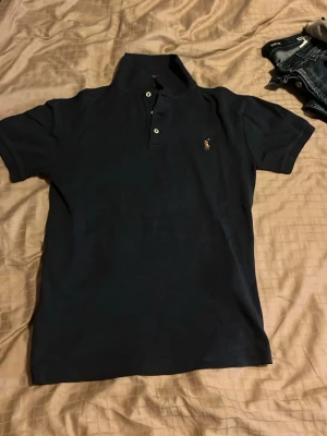 Svart pikétröja från Ralph Lauren - Klassisk svart pikétröja från Ralph Lauren med korta ärmar och två knappar framtill. Tröjan har den ikoniska broderade loggan på bröstet och är tillverkad i mjuk bomull. Perfekt till jeans eller shorts för en clean look. Denna piké är perfekt inför sommaren och för ett riktigt bra pris eftersom den är sjukt bra skick och har använts ett fåtal gånger. Säljer den endast för 349kr och nypris är 1095. Säljs för 349 bara eftersom jag inte har användning av den och vill ha något för den.