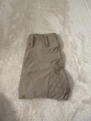 Beige ridbyxor med helskodd - Snygga beige ridbyxor med helskodd i silikonmönster för extra grepp. Byxorna har en bakficka med knapp, bälteshällor och diskret logga på benet. Perfekta för träning eller tävling inom ridsport. Materialet är stretchigt och följsamt för bästa komfort. Från maya delorez 