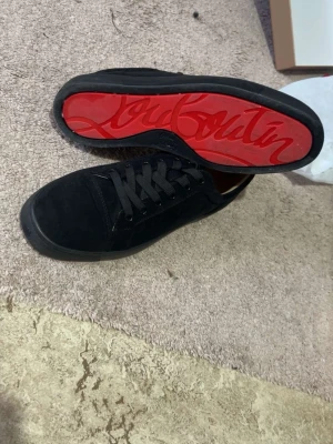 Svarta sneakers från Christian Louboutin - Stilrena svarta sneakers i mocka från Christian Louboutin med klassisk röd sula och signaturdetaljer undertill. Skorna har svarta skosnören och låg profil som ger en clean look. Perfekta för dig som vill ha något exklusivt och tidlöst. Använt endast 2 tillfällen 