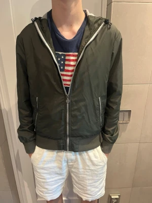 Olivgrön vindjacka från woolrich - Snygg olivgrön vindjacka med dragkedja och huva. Jackan har ribbade muddar vid ärmslut och midja samt två fickor framtill. Justerbar dragsko i huvan och lätt material som passar perfekt till vår och sommar.