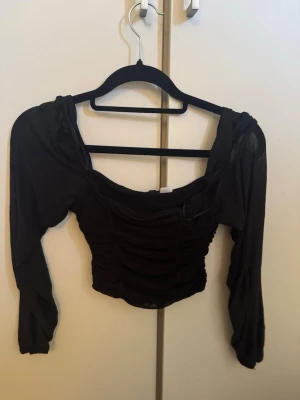 Svart croppad topp med rynk - Trendig svart croppad topp med långa ärmar och rynkade detaljer framtill. Toppen har en bred, fyrkantig urringning och är tillverkad i ett stretchigt material som sitter snyggt på kroppen. Perfekt för dig som gillar en edgy och modern stil.