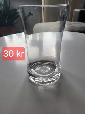 Vas  - Säljer ett stilrent, transparent vas med lätt utsvängd form och tjock botten. Perfekt som en enkel inredningsdetalj. Vasen är helt genomskinligt och har en modern look som passar i de flesta hem.
