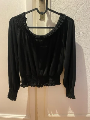 Svart blus med volang från Gina Tricot - Supersöt svart blus från Gina Tricot med volangdetaljer vid halsringning, ärmslut och midja. Blusen har långärmade puffärmar och smockdetaljer som ger en snygg passform. Perfekt att styla med jeans eller kjol för en trendig look. Använd några gånger i riktigt bra skick. 