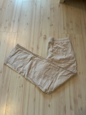 Beiga linnebyxor - Säljer ett par beiga linnebyxor med avslappnad passform och resår i midjan samt dragsko. Byxorna har raka ben, sidofickor och en bakficka. Perfekta för en chill och stilren look under varma dagar.