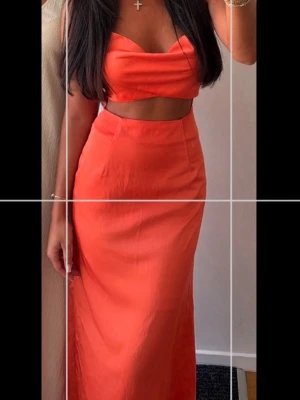Orange klänning med crop top-look - Snygg orange klänning med trendig crop top-design och hög midja. Klänningen har en figurnära passform och faller slätt, perfekt för dig som vill sticka ut. Materialet är satin  mjukt och följsamt, vilket ger en skön känsla. Inga  fickor eller detaljer.