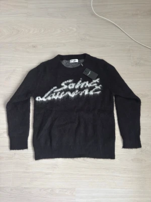Svart Saint Laurent stickad tröja - Stickad svart tröja från Saint Laurent med vit logotyp i borstigt garn framtill. Tröjan har rund halsringning, långa ärmar och ribbade muddar. Materialet ser ut att vara en mjuk och fluffig ullblandning som ger en lyxig känsla.