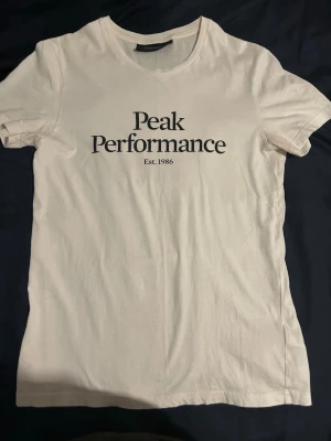 Vit Peak Performance t-shirt - Klassisk vit t-shirt från Peak Performance med svart logotyptryck på bröstet. T-shirten har rund halsringning och korta ärmar. Tillverkad i mjuk bomull för en skön känsla och enkel stil.