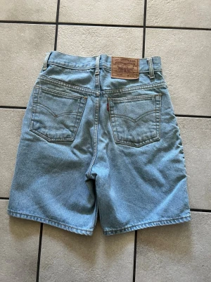 Levi's 501 ljusblå jeansshorts storlek 27 - Klassiska ljusblå jeansshorts från Levi's modell 501 med hög midja och raka ben. Shortsen har två bakfickor och den ikoniska läderpatchen bak i midjan. Perfekta för en avslappnad och trendig stil. Midjemått 70 cm, längd 48 cm