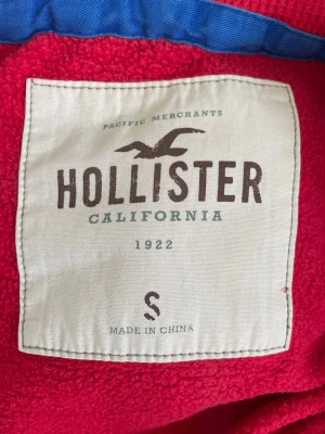 Röd hoodie från Hollister - Snygg röd hoodie från Hollister med klassisk huva och mjukt material. Hoodien har en relaxed fit och ribbade muddar vid ärmslut och nederkant. Perfekt för dig som gillar en enkel men stilren look. Finns några skavanker som några fläckar och lite trasig i mudden men absolut inget som syns om du skulle stå mittemot någon.