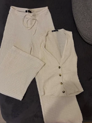 Krämvitt ribbat set från Zara - Säljer ett krämvitt ribbat set från Zara med byxa och ärmlös body. Setet har en mjuk struktur, knappar framtill på bodyn och byxorna har knytband i midjan. Perfekt för en avslappnad och trendig look.