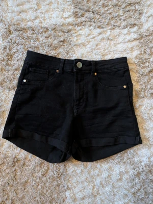 Svarta denimshorts med uppvik - Snygga svarta shorts i denim med klassisk femficksmodell och uppvikta benslut. De har knapp och dragkedja framtill samt bälteshällor. Perfekta för sommaren och passar till det mesta.
