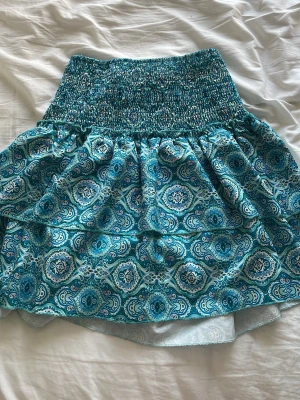 Turkos mönstrad kortkjol - Säljer en turkos kortkjol med elastisk smock i midjan och ett snyggt paisleyinspirerat mönster i blått och vitt. Kjolen har en luftig passform och är perfekt för varma dagar. Materialet känns lätt och bekvämt.