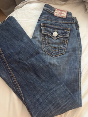 Blå True Religion jeans med kontrastsömmar - Säljer ett par klassiska blå jeans från True Religion med tydliga kontrastsömmar och ikonisk fickdesign bak. Jeansen har raka ben, låg midja och detaljerade knappar. Materialet är slitstarkt denim i bomull. Perfekta för dig som gillar streetwear och vill sticka ut. Ett litet litet hål är vid undersidan av rumpan, den kan fixas enkelt, säljer då dom ej passar mig längre. OBS dom är köpta secondhand 