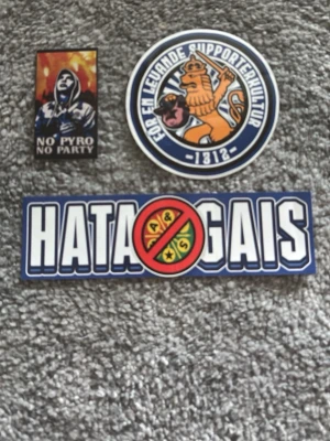 IFK Göteborg supporterklistermärken - Tre olika klistermärken för IFK Göteborg-supportrar. Ett runt med lejon och texten 'För en levande supporterkultur', ett rektangulärt med motiv av en person och texten 'No pyro no party', samt ett avlångt med texten 'Hata Gais' och överstruket GAIS-emblem.
