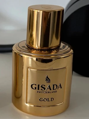 Gisada Gold parfym - Lyxig parfymflaska från Gisada Switzerland i en glänsande guldfärgad design. Flaskan är rund med en stor, matchande guldfärgad kork och har en elegant etikett med svart text. Perfekt för dig som gillar exklusiva dofter och snygg inredningsdetalj. Köptes ny för 800kr, endast testad