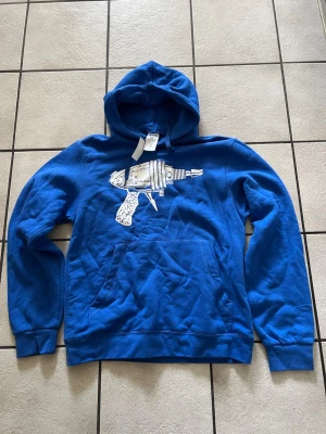 Blå hoodie med raygun-tryck storlek S 80% bomul - Snygg blå hoodie med ett stort vitt raygun-tryck på bröstet. Hoodien har klassisk känguruficka och dragsko i huvan. Tillverkad i mjukt bomullsmaterial som känns skönt mot huden. Perfekt för dig som gillar coola och unika prints.