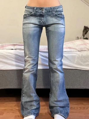 Lowaist bootcut jeans med slitningar - Snygga blå bootcut jeans med lätt slitna detaljer och klassisk femficksdesign. Jeansen har låg midja och utställda ben, vilket ger en retro vibe. Tillverkade i denim med ljus tvätt och kontrastsömmar. Perfekta för dig som gillar en avslappnad men trendig stil.