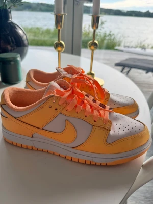 Nike Dunk Low orange och vita sneakers - Fräscha Nike Dunk Low sneakers i orange och vitt med klassisk swoosh på sidan. Skorna har lågt skaft, perforerad tåbox och matchande orange skosnören. Ovandelen är i läder och mesh för en snygg och sportig look. Perfekta för dig som gillar färg och streetstyle.
